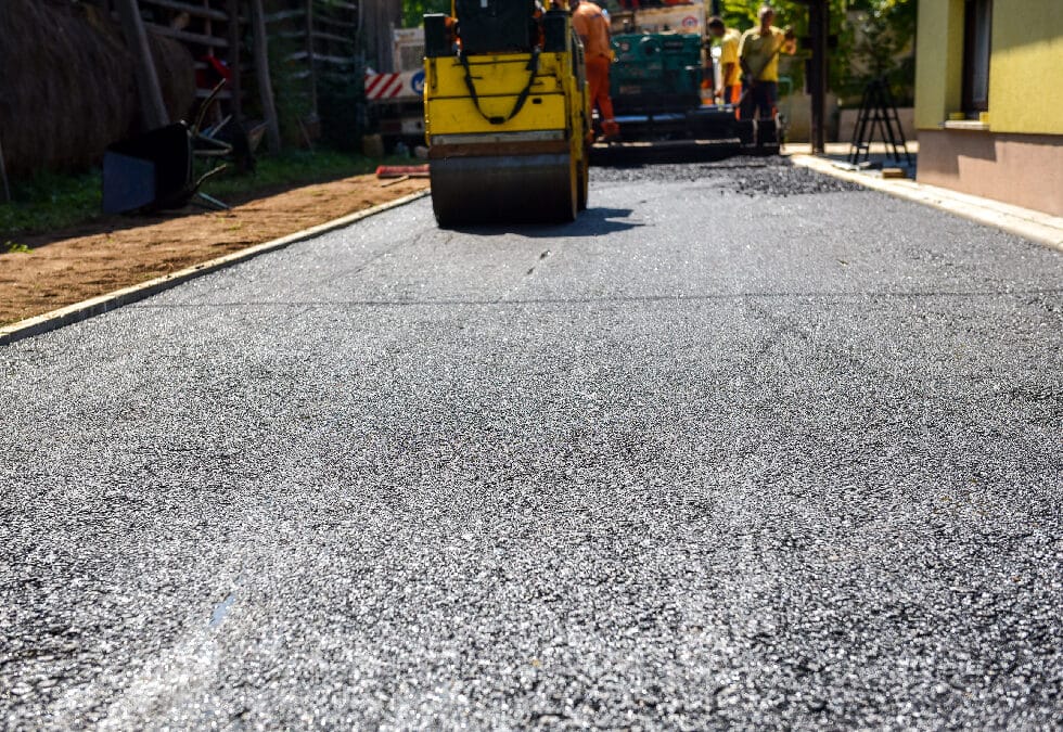 A low-angle, close-up view focusing on the rough texture of freshly laid black asphalt. In the blurred background, a heavy yellow road roller drives away from the camera, compacting the new pavement, while a construction crew works with paving machinery on a sunny day.