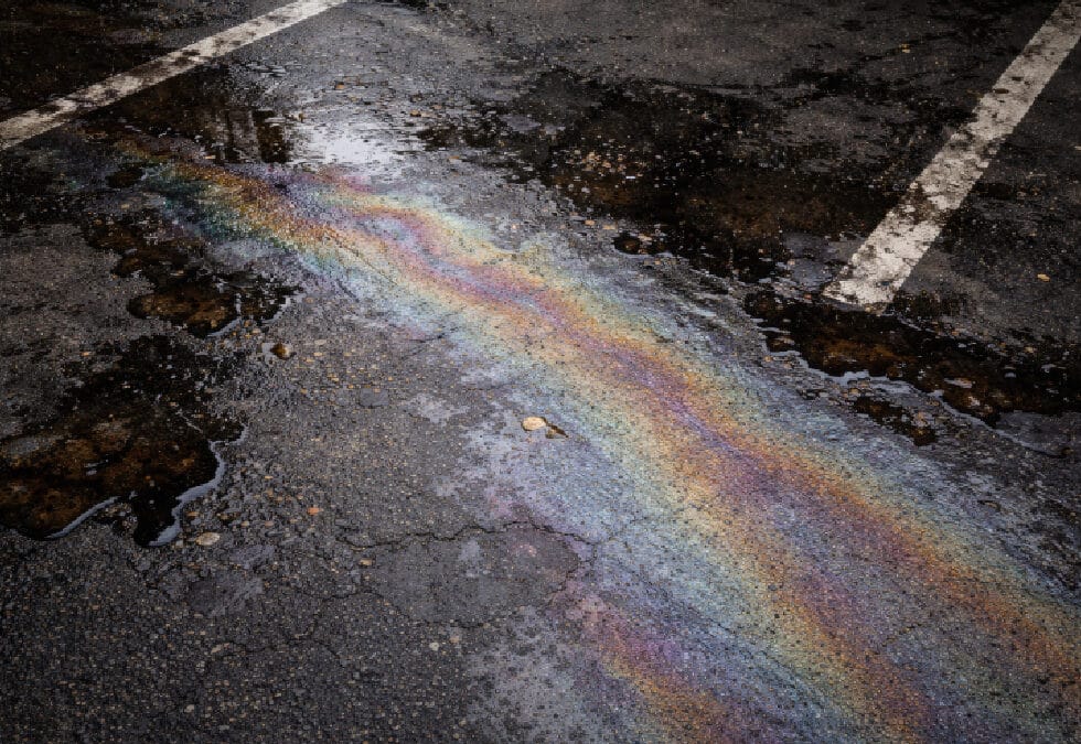 A high-angle shot of a vibrant, iridescent rainbow-colored oil sheen spreading across wet, cracked asphalt pavement. The colorful fuel slick stretches diagonally between faded white painted lines of a parking space, mixed with dark puddles of water.